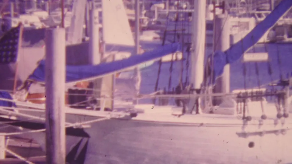 1964 Honolulu harbor frame centered on a white sailboat with mast, rigging, blue canvas cover, dock lines, and moored boats behind it.