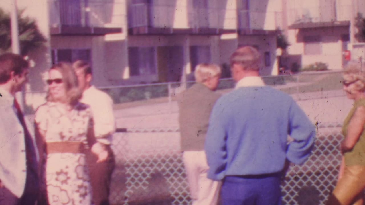 1964 Honolulu home-movie frame showing adults in 1960s clothing conversing near a chain-link fence with an apartment building behind them.