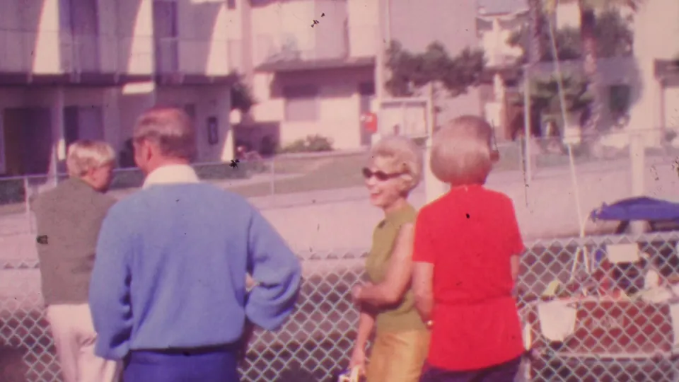 1964 Honolulu outdoor home-movie frame with adults in casual mid-century clothing standing by a chain-link fence and apartment exterior.