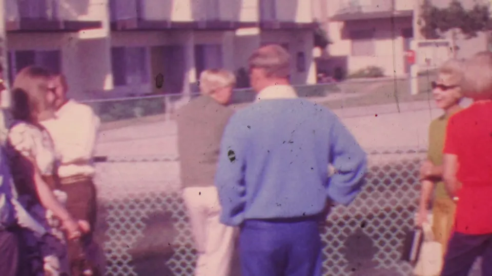 1964 Honolulu frame of adults in period clothing standing and talking near a chain-link fence, with the camera catching the group from a casual home-movie angle.