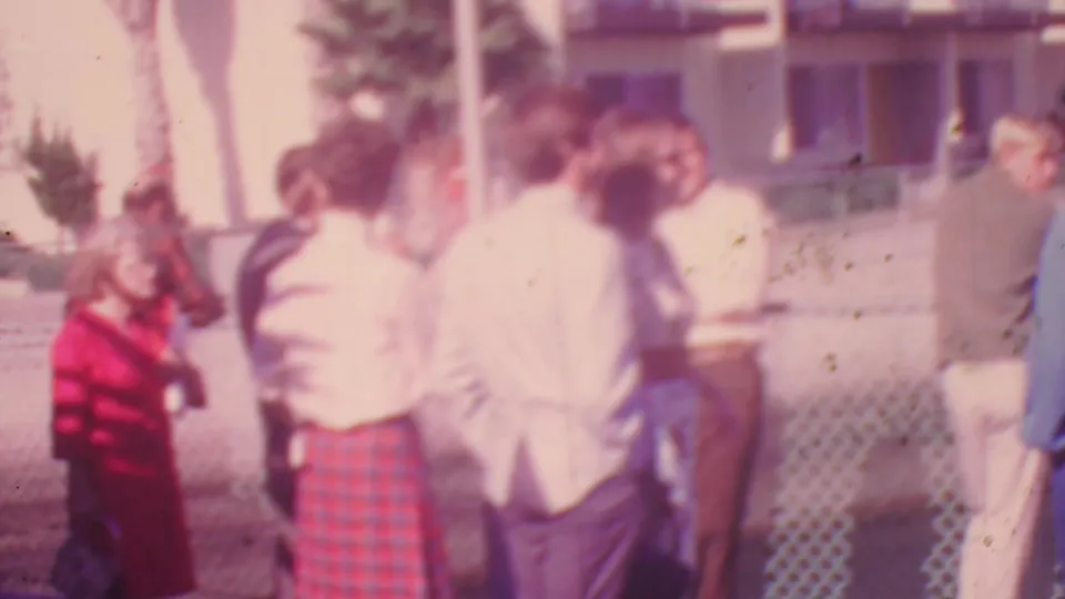 Soft 1964 Honolulu home-movie frame showing adults near a chain-link fence, including a woman in a red plaid skirt and men in light shirts.