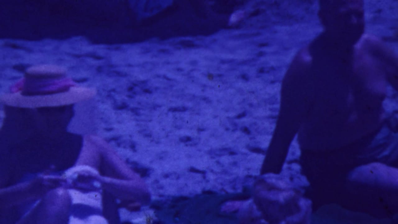 Adults relaxing on sandy ground with beach chairs and a straw hat visible under a blue-purple color cast.