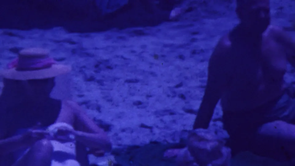 1964 Palm Springs home-movie frame showing adults relaxing on sand, with a seated figure in a straw hat and nearby beach chairs under a strong blue-purple 8mm color shift.