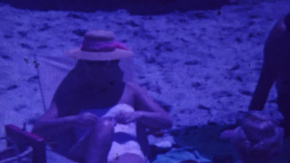 1964 Palm Springs frame with a hat-wearing adult reclining in a folding beach chair and another adult seated on sand, rendered with soft 8mm grain and blue-purple color.