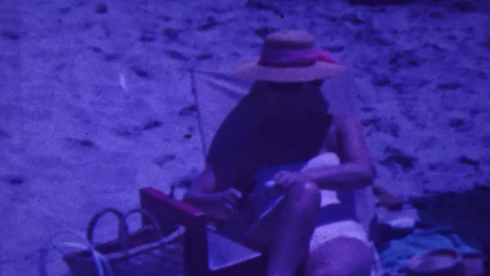 1964 Palm Springs home-movie still showing an adult in a straw hat seated in a folding beach chair, with sand, bags or towels, and swimwear details visible.