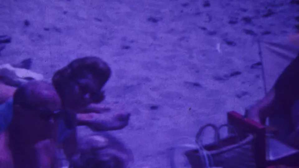1964 Palm Springs frame showing several adults gathered on sandy ground near folding beach chairs and beach items, with partially visible faces and pronounced blue-purple 8mm coloration.