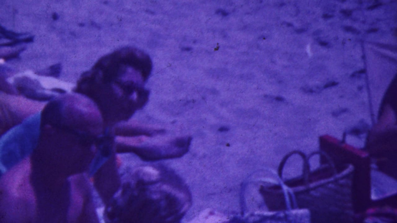 1964 Palm Springs home-movie frame showing adults in swimsuits sitting and reclining on a sandy beach area with towels and bags nearby.