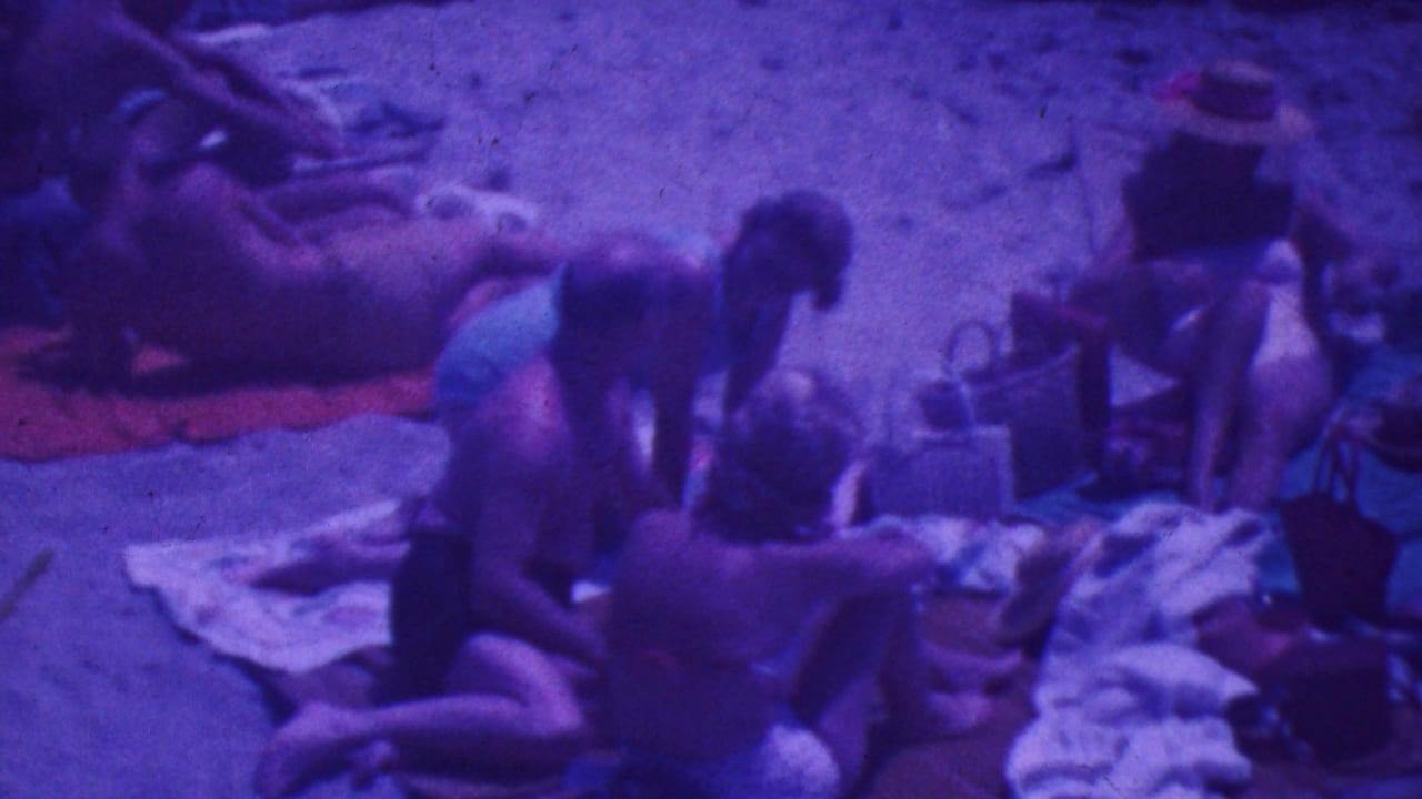 1964 Palm Springs beach scene with adults in period swimwear seated on towels and one person lying nearby, captured with a strong purple 8mm color shift.