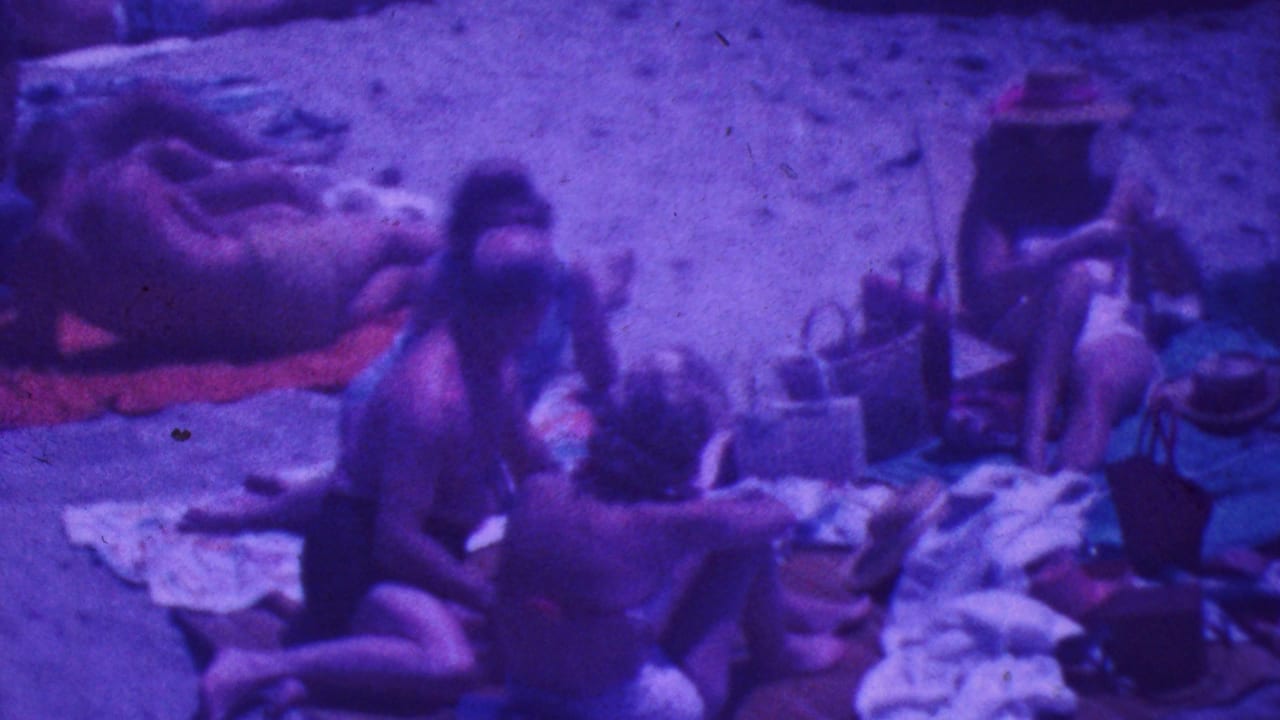 Beach group gathers around towels and bags while a woman in a sunhat sits nearby.