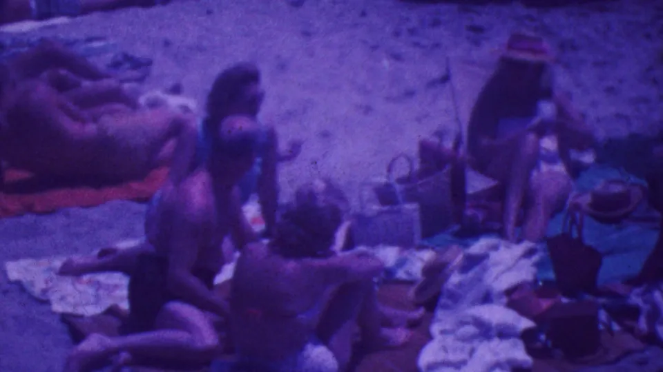1964 Palm Springs beach footage frame with adults seated and reclining on sand beside towels and bags, with a sunhat-wearing figure at the right edge.