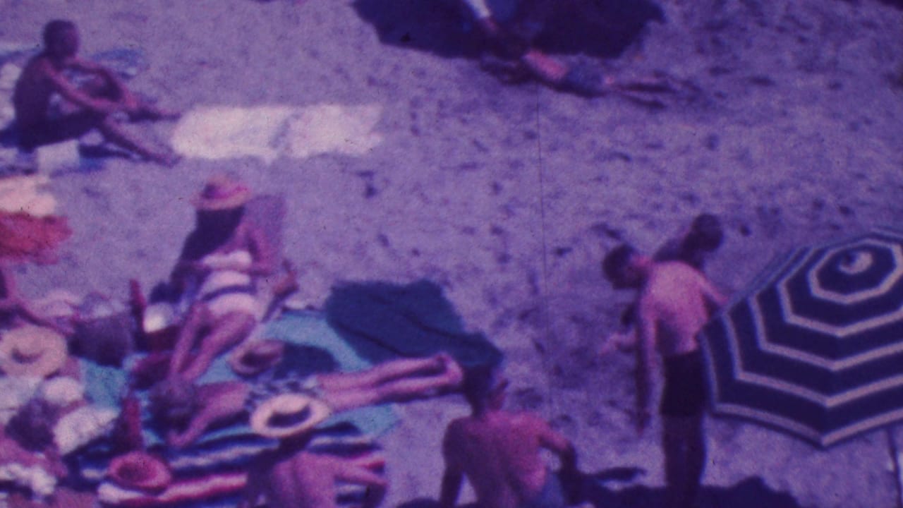 Adults relaxing on a sandy beach with towels, hats, and a striped umbrella nearby.
