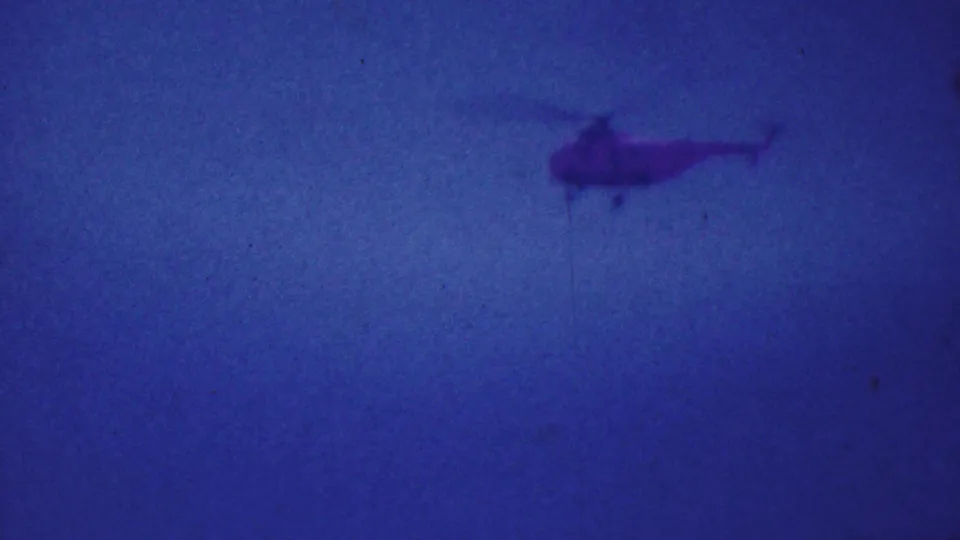 Soft, grainy 8mm frame of a distant helicopter at the edge of a deep blue sky, from the 1964 Palm Springs home-movie clip.