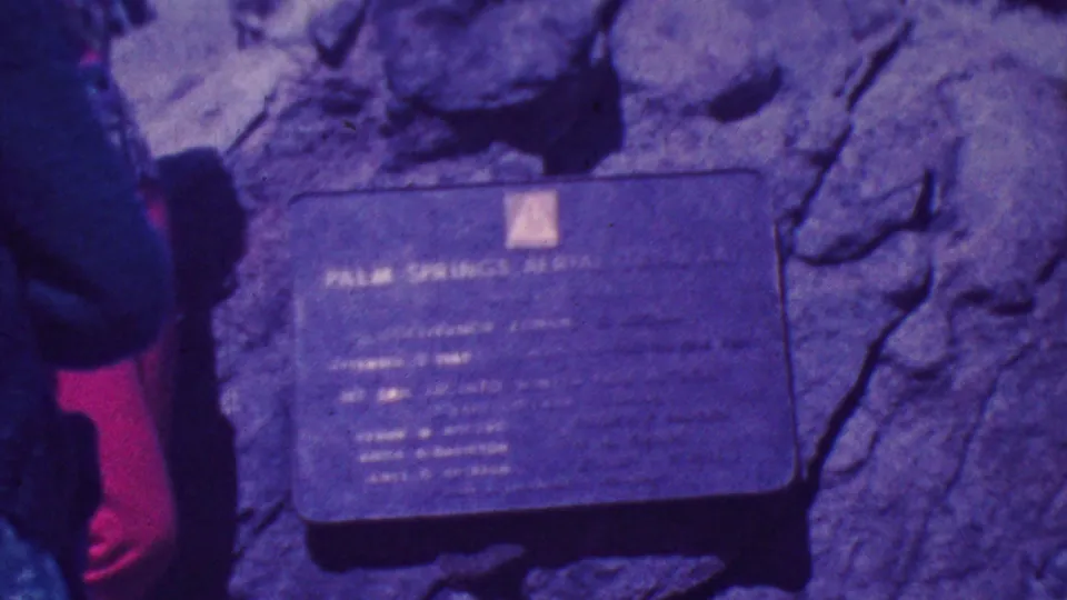 The frame centers on a metal plaque set against rough rock, with partial adult figures at the edges and the aged 8mm color cast making the text difficult to read.