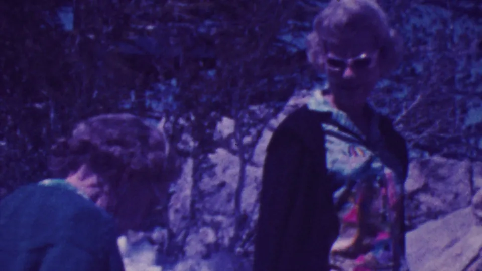 Two adult visitors are visible in the rocky Palm Springs setting, including a sunglasses-wearing woman in a patterned top and another person bending or looking downward near the stones and vegetation.