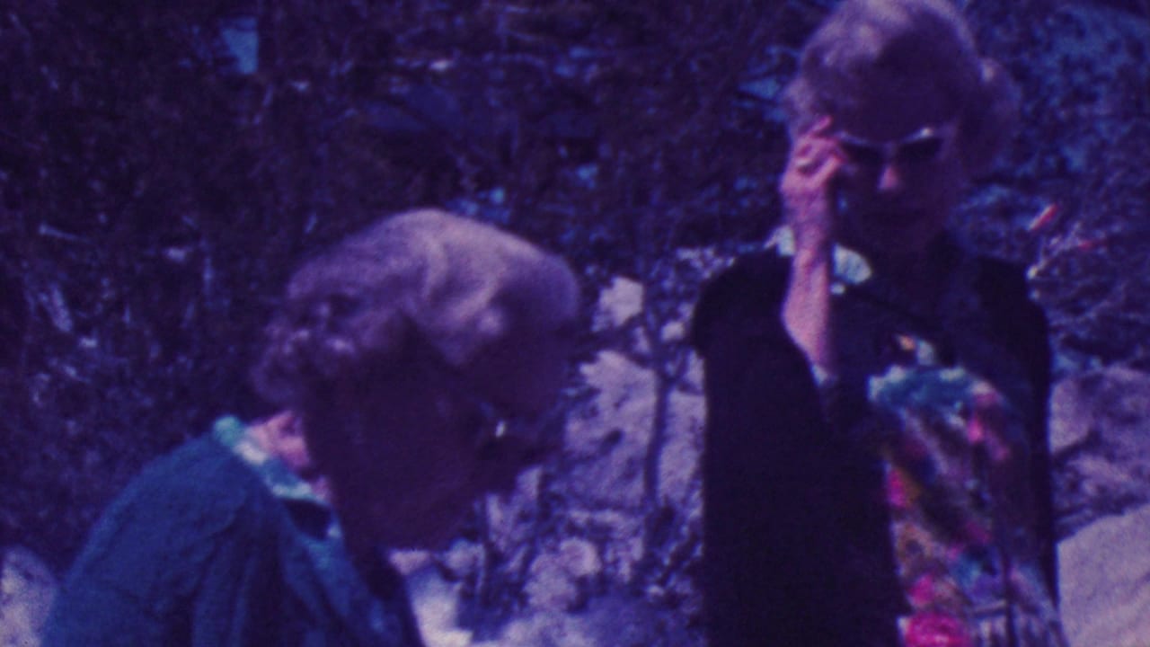 Older woman in profile wearing sunglasses stands near another woman in a dark cardigan among trees.