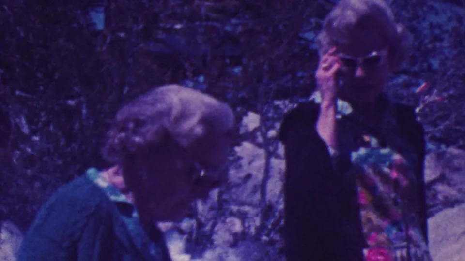 1964 Palm Springs home-movie frame with an older woman in sunglasses seen in profile beside another woman in a wooded rocky outdoor area.