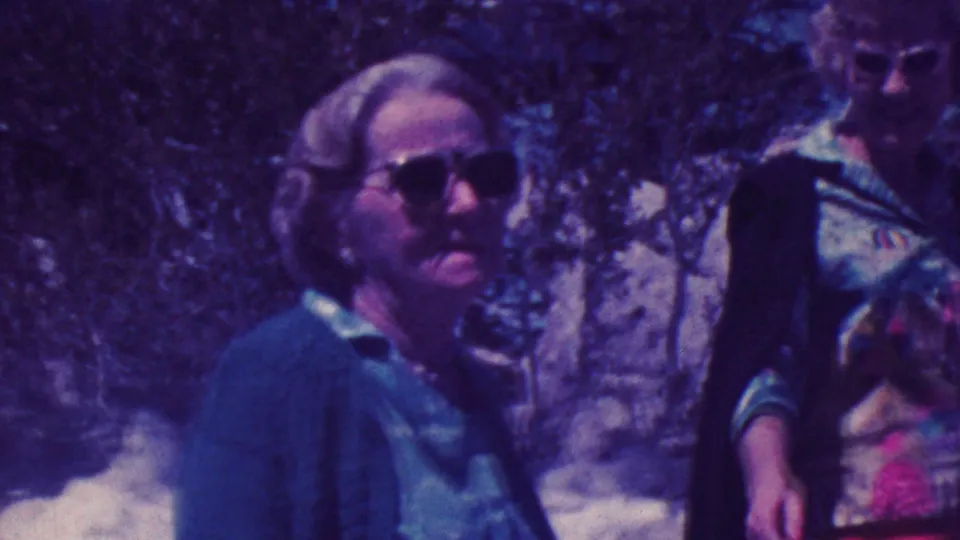 1964 Palm Springs home-movie frame showing three older women close to the camera in a rocky wooded setting, softened by grain and purple color shift.