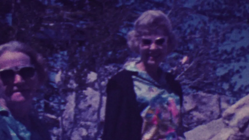 1964 Palm Springs home-movie frame showing an older woman in a dark cardigan and patterned blouse standing among trees and pale rocks.