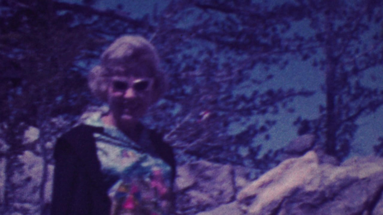 A blonde woman wearing sunglasses, a floral top, and a dark sweater stands near rocks and trees in 1964 Palm Springs home-movie footage.