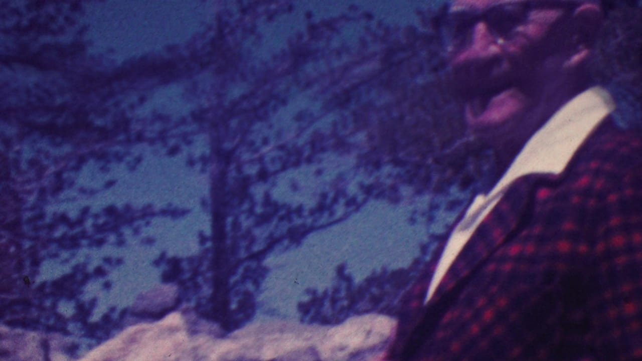 An older bald man wearing glasses and a red plaid jacket smiles toward the camera in outdoor Palm Springs vacation footage from 1964.