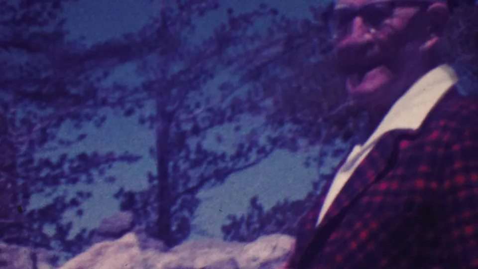 An older bald man wearing glasses and a red plaid jacket smiles toward the camera in outdoor Palm Springs vacation footage from 1964.