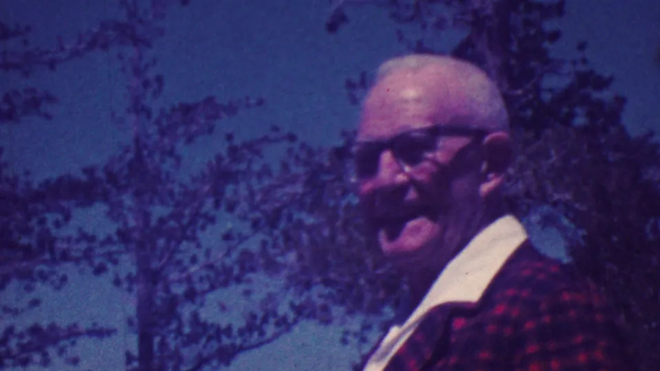 The older man with glasses and a red plaid jacket is seen closer in frame, with trees and sky behind him in purple-shifted 8mm footage.