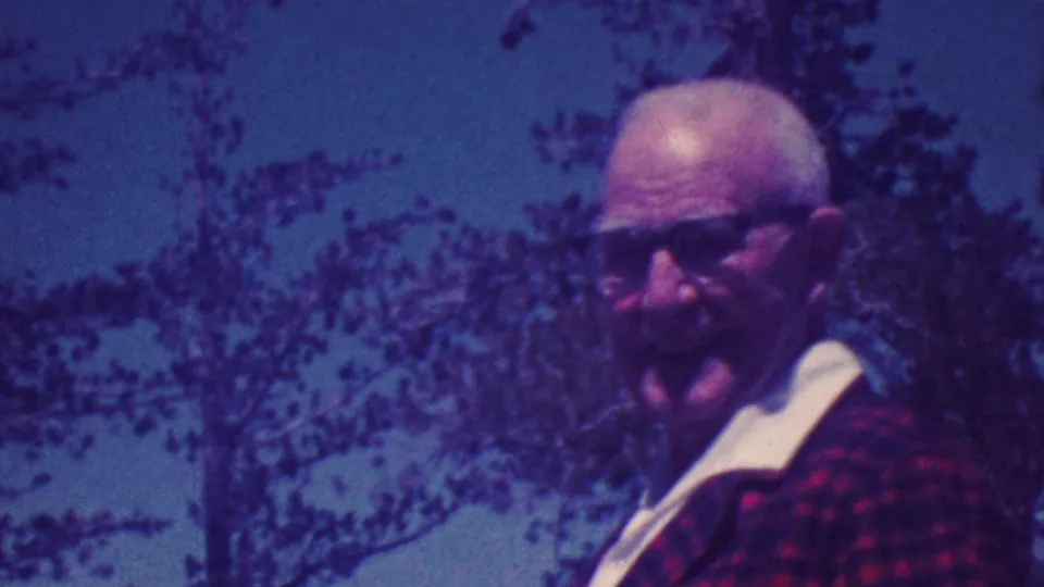 An older man in glasses and a red plaid jacket looks toward the camera with trees and sky behind him in the 1964 Palm Springs vacation clip.