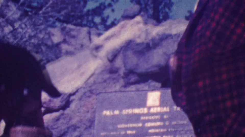A weathered memorial plaque set among rocks appears near a partial adult figure, with foliage and rugged outdoor terrain visible in the frame.