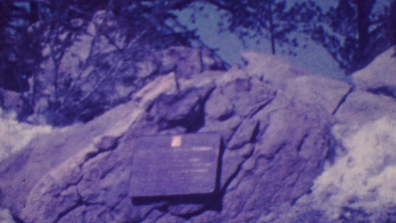 1964 Palm Springs-area 8mm frame showing a dark memorial plaque fixed to a large boulder, with snow patches and trees in the background.