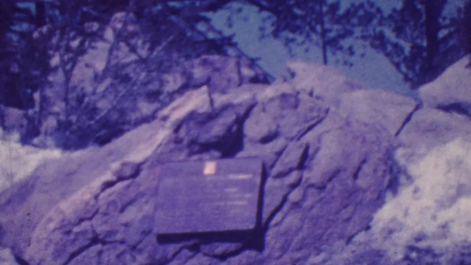1964 Palm Springs-area 8mm frame showing a dark memorial plaque fixed to a large boulder, with snow patches and trees in the background.