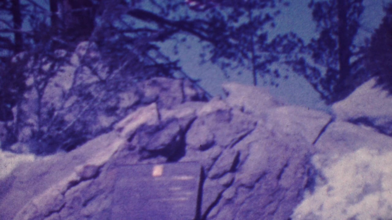 1964 Palm Springs-area frame with the memorial plaque still visible on the rock as the camera rises toward a small American flag among trees.