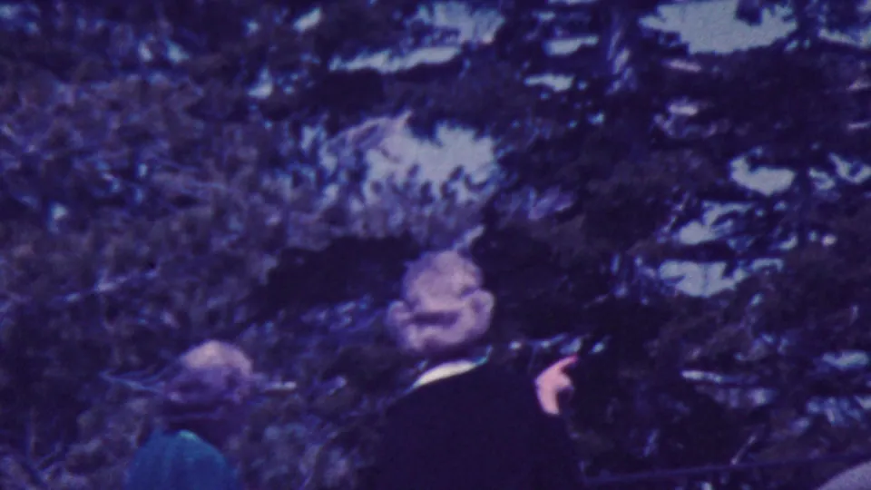 1964 Palm Springs home-movie frame showing adults partly obscured by evergreen trees, with one person in a dark jacket turning near the camera.