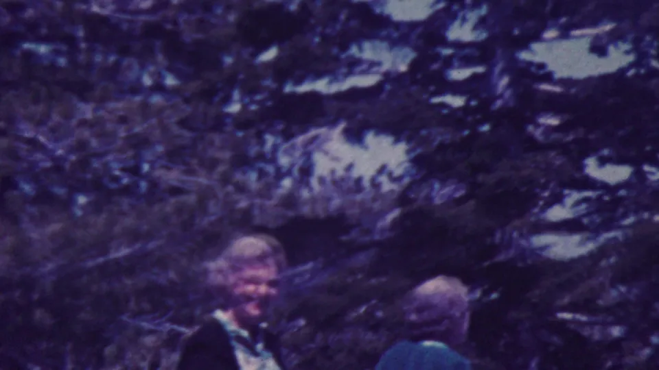 1964 Palm Springs home-movie frame showing two adults near evergreen branches, including a smiling person in dark clothing facing the camera.