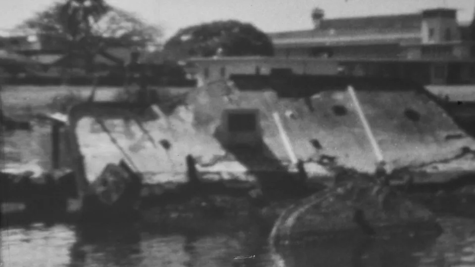1962 Honolulu/Pearl Harbor-area frame showing a damaged structure partly surrounded by floodwater, with debris and an elevated bridge or walkway crossing the background.