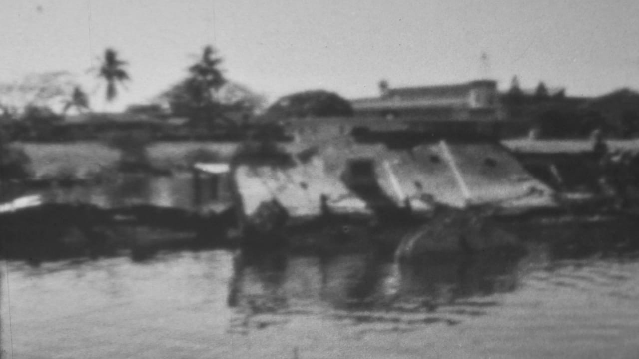 High water and damaged shoreline structures appear in front of low buildings and trees.