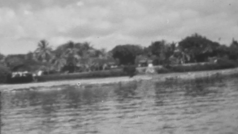 Black-and-white 1962 8mm frame showing calm Pearl Harbor-area water with a palm-lined shoreline and low waterfront structures in the distance.