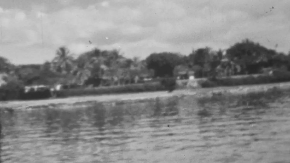 Black-and-white 1962 8mm frame from the Pearl Harbor, Honolulu area with low shoreline houses, palms, and rippled harbor reflections.