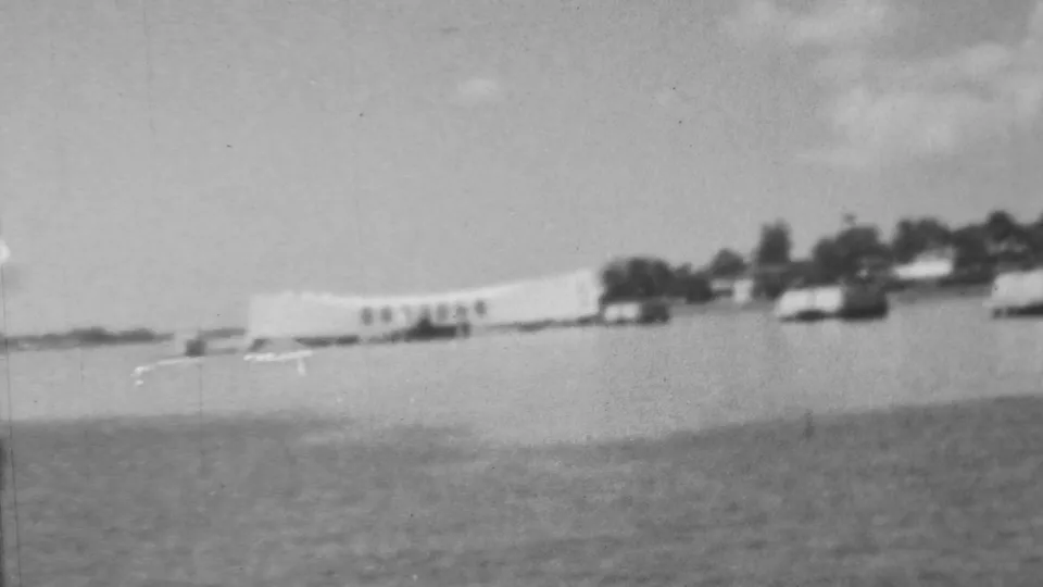 Black-and-white 1962 8mm frame showing a wider Pearl Harbor-area water view with distant shoreline structures and small boats or waterfront shapes.