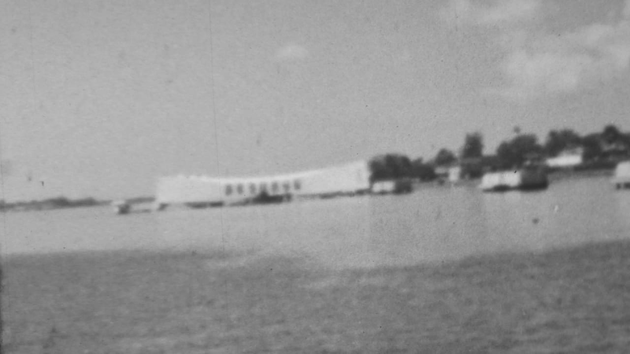 Distant 1962 home-movie view of the Pearl Harbor memorial area in Honolulu, seen across broad harbor water with small boats and a low shoreline nearby.