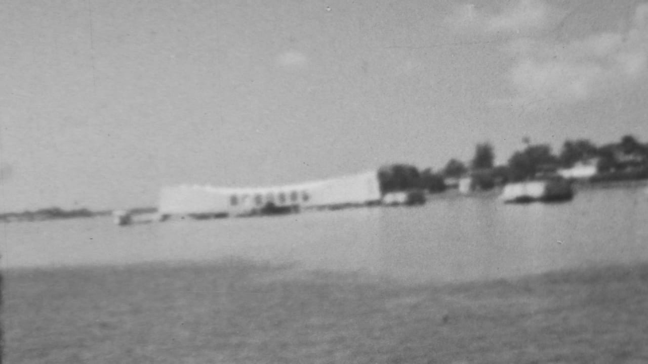 Soft black-and-white 8mm frame showing the Pearl Harbor memorial area from across the water, with small boats near the shoreline and trees in the background.