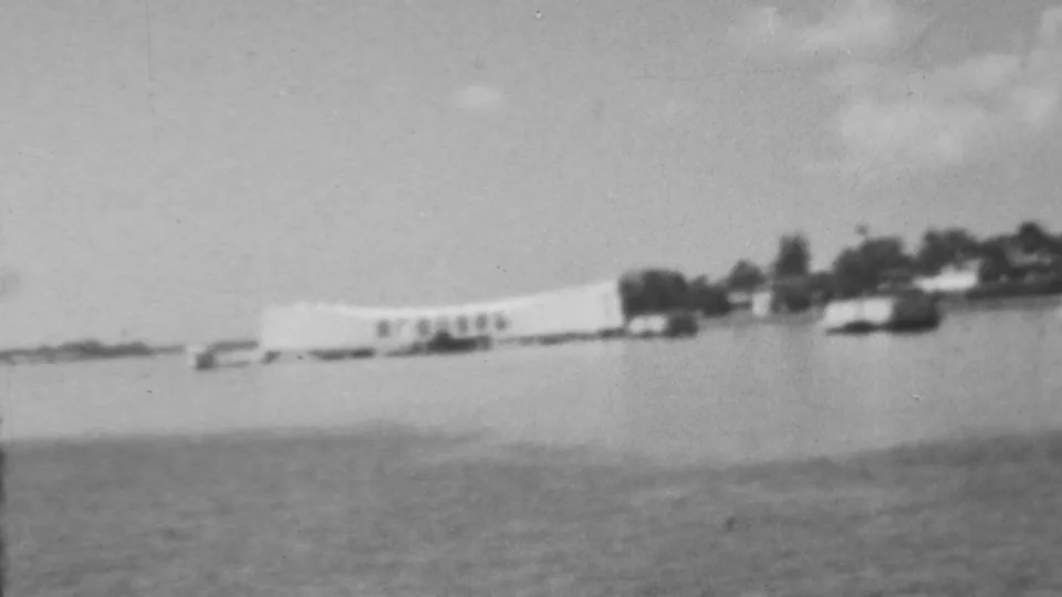 Soft black-and-white 8mm frame showing the Pearl Harbor memorial area from across the water, with small boats near the shoreline and trees in the background.