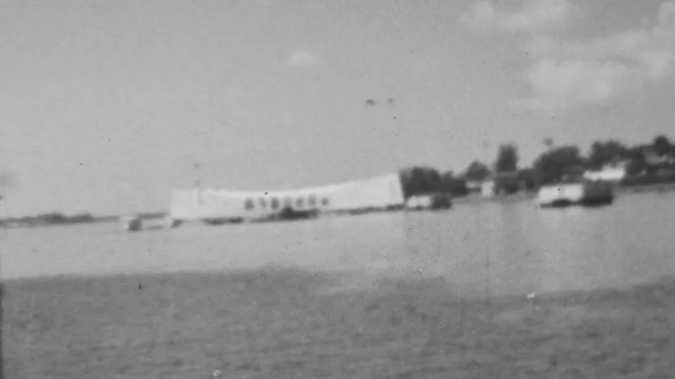 1962 8mm home-movie frame of the Pearl Harbor memorial area from a low, distant vantage point across the harbor, with boats and shoreline details softened by film grain.