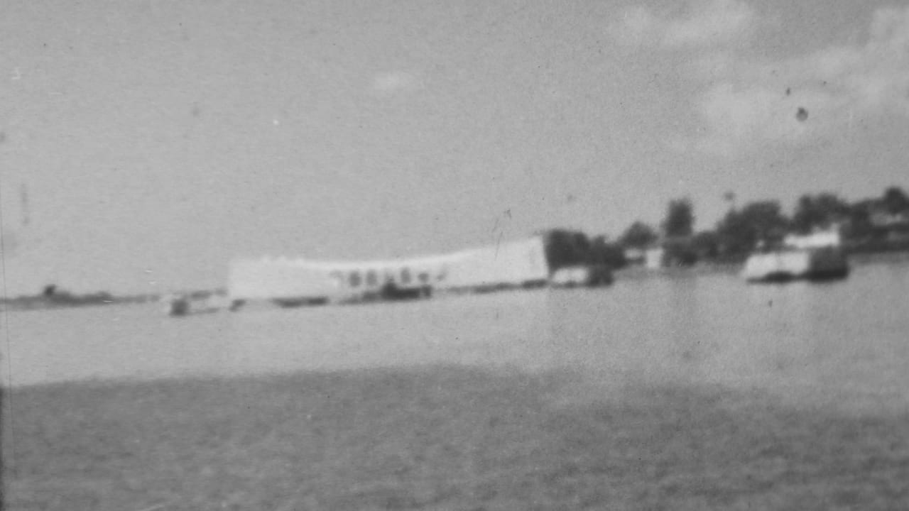 Black-and-white 1962 8mm view of the USS Arizona Memorial at Pearl Harbor seen from across the water, with small boats and shoreline trees in the distance.