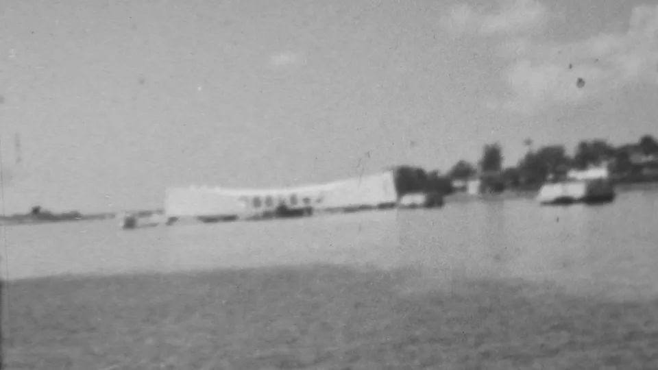 Black-and-white 1962 8mm view of the USS Arizona Memorial at Pearl Harbor seen from across the water, with small boats and shoreline trees in the distance.