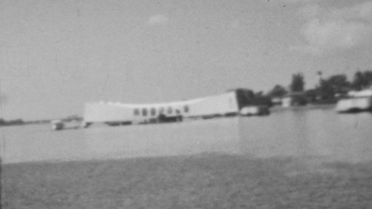 The USS Arizona Memorial appears in soft black-and-white focus across Pearl Harbor water, with a small boat and tree-lined shoreline behind the structure.