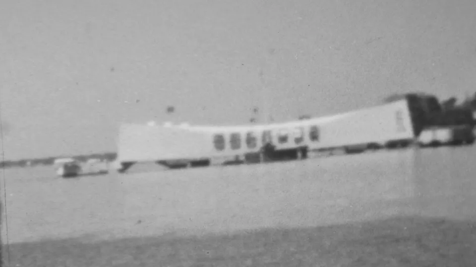 Soft black-and-white 8mm frame of the USS Arizona Memorial at Pearl Harbor, showing the long white form, dark window openings, and calm water in the foreground.