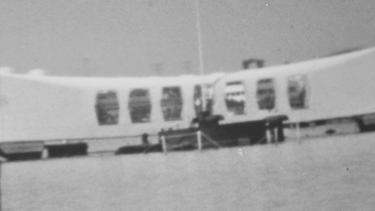 1962 black-and-white 8mm frame showing the USS Arizona Memorial at Pearl Harbor from a visitor viewpoint across the water, with arched windows and a dark structure below.