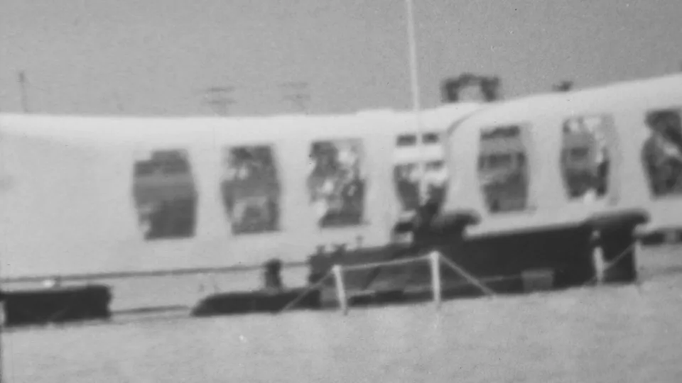 1962 Pearl Harbor home-movie frame showing the USS Arizona Memorial in soft focus, with arched windows, a railing, dark dock-like forms, and harbor water in the foreground.