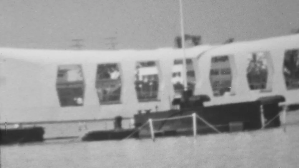 Black-and-white 8mm frame from Pearl Harbor showing the USS Arizona Memorial, its arched window openings, a flagpole, railings, and calm water in the foreground.