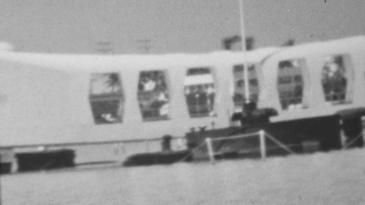 1962 black-and-white 8mm frame showing the curved Arizona Memorial at Pearl Harbor across the water, with open window bays, railings, and small visitor figures near the structure.